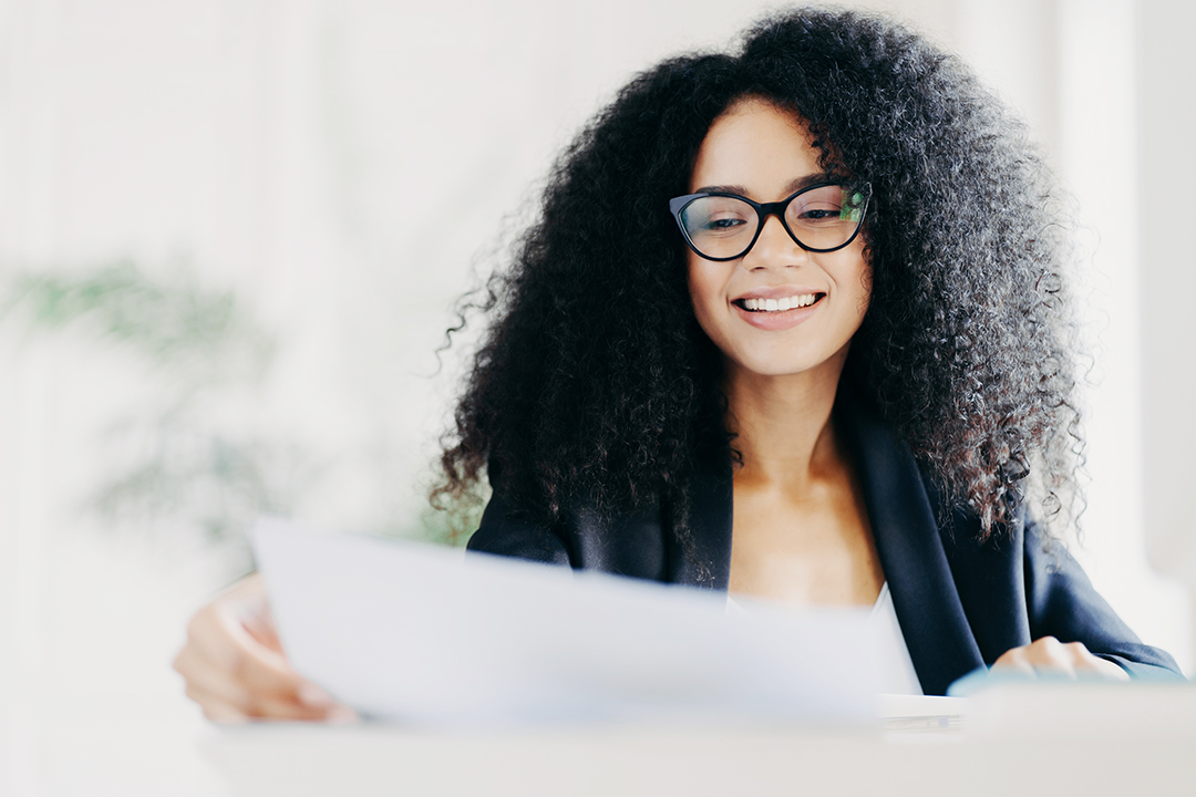 Happy Afro American woman with curly hairstyle, looks through documents, prepares financial report, wears transparent glasses and formal suit, poses in her cabinet, works on financial report